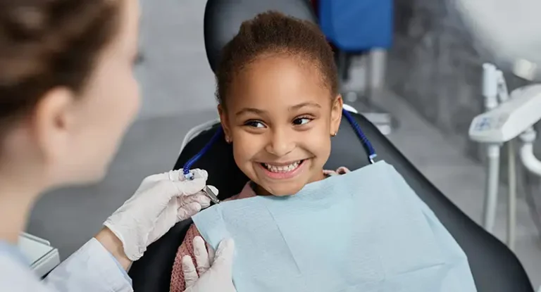 Pediatric dentistry: a smiling girl has a bib placed on her by a dental assistant.