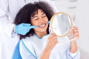 A smiling patient lying on a dental chair uses a mirror to look at her teeth whitening results.