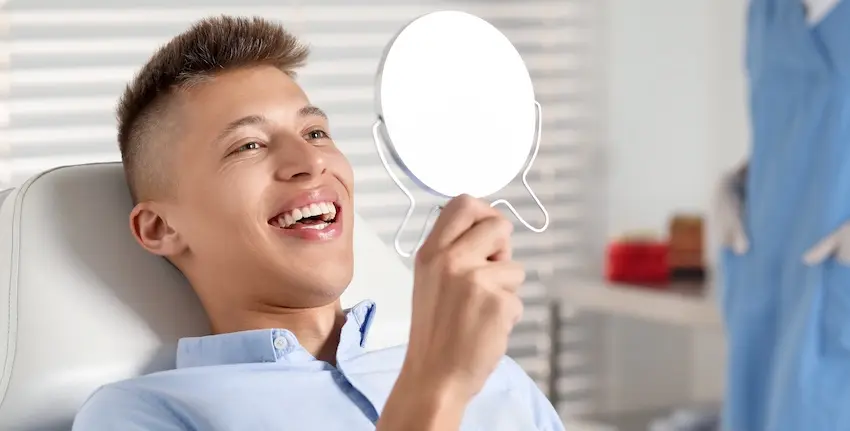A happy young man is smiling in a dental chair and is holding a mirror while admiring the successful dental implants that he had just received at The Foleck Center in the Tidewater Virginia area.