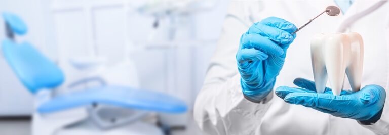 A dentist in blue gloves holds a large tooth model and a dental mirror tool in front of a blurred dental chair in a bright clinical office at The Foleck Center representing the professional tools and environment used to perform a thorough dental cleaning.