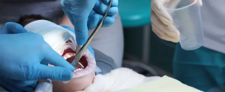A dentist in blue gloves uses a dental instrument inside a reclined patient's open mouth while an assistant stands nearby depicting the precise clinical procedure involved in placing a dental filling while under anesthesia at The Foleck Center.