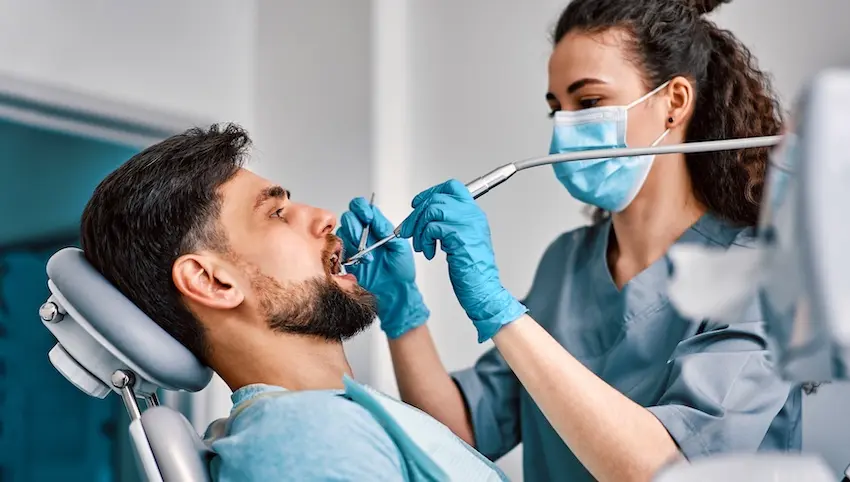 A female dentist wearing a surgical mask and blue gloves works inside a bearded male patient's mouth using a drill and suction tool showing the step-by-step process a patient experiences when receiving a dental filling at The Foleck Center in Virginia.