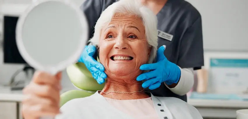 An elderly woman smiles brightly at her reflection in a hand mirror while a gloved dental professional gently frames her face expressing the confidence and satisfaction patients feel after completing a dental filling and smile restoration in Virginia.