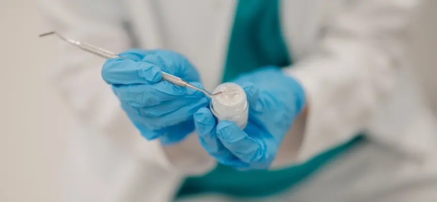 A close-up of a gloved dentist's hands holding a metal dental probe and a small container of composite material capturing the tools and bonding agent used to prepare and place a dental filling at The Foleck Center in the Tidewater Virginia area.