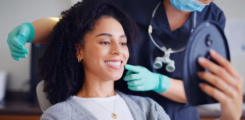 A smiling woman with natural black curly hair admires her teeth in a handheld mirror while a dentist in teal gloves and a mask points to her smile depicting the confident results a patient can expect after a professional dental cleaning at The Foleck Center in Tidewater Virginia.