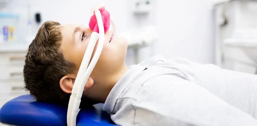 A young boy reclines in a dental chair with a pink nitrous oxide mask over his nose representing the use of sedation to keep anxious pediatric patients calm and comfortable during a dental cleaning at The Foleck Center.