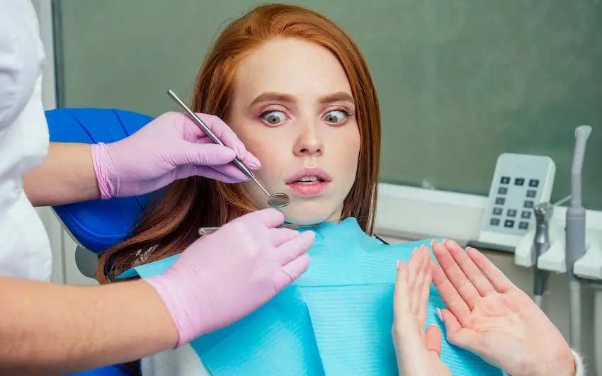 A wide-eyed young woman in a dental chair raises her hand as if to stop the procedure while a dentist in pink gloves approaches with a dental mirror vividly depicting the panic and distress experienced by patients suffering from dental phobia in Virginia.