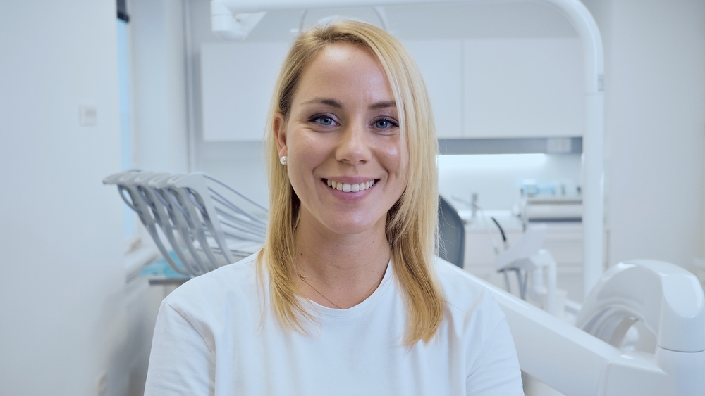 A smiling female dentist in a modern, clean dental office. She wears a white uniform, exuding professionalism and warmth.