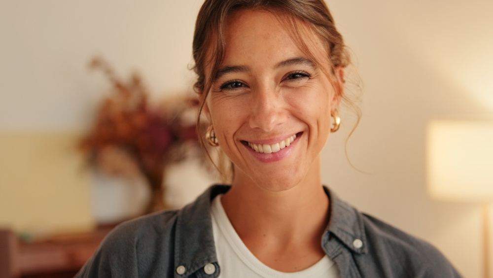 A warm, friendly portrait of a smiling woman in a casual denim shirt with gold hoop earrings, framed by soft home lighting for a welcoming, lifestyle vibe.