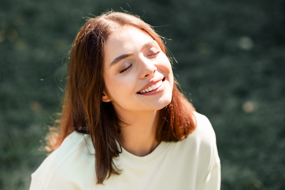 A young woman with closed eyes smiles peacefully as warm sunlight hits her face in a natural outdoor portrait, conveying relaxation, joy, and calm on a sunny day.