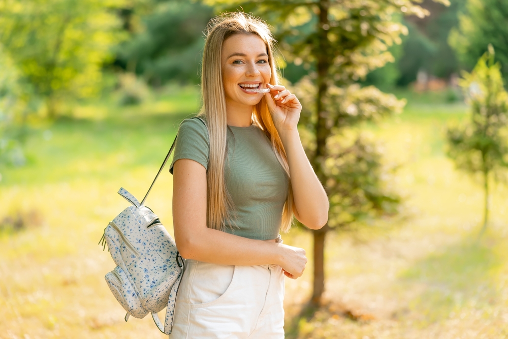 Retainer Use Outdoors: Virginia Beach Full-Mouth Reconstruction Patient Young woman placing a clear dental retainer in her mouth outdoors after full-mouth reconstruction in Virginia Beach
