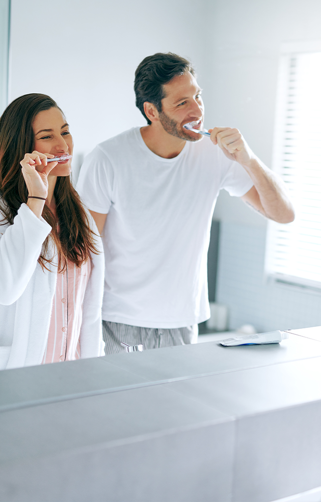 Man and woman brushing their teeth together in front of a bathroom mirror