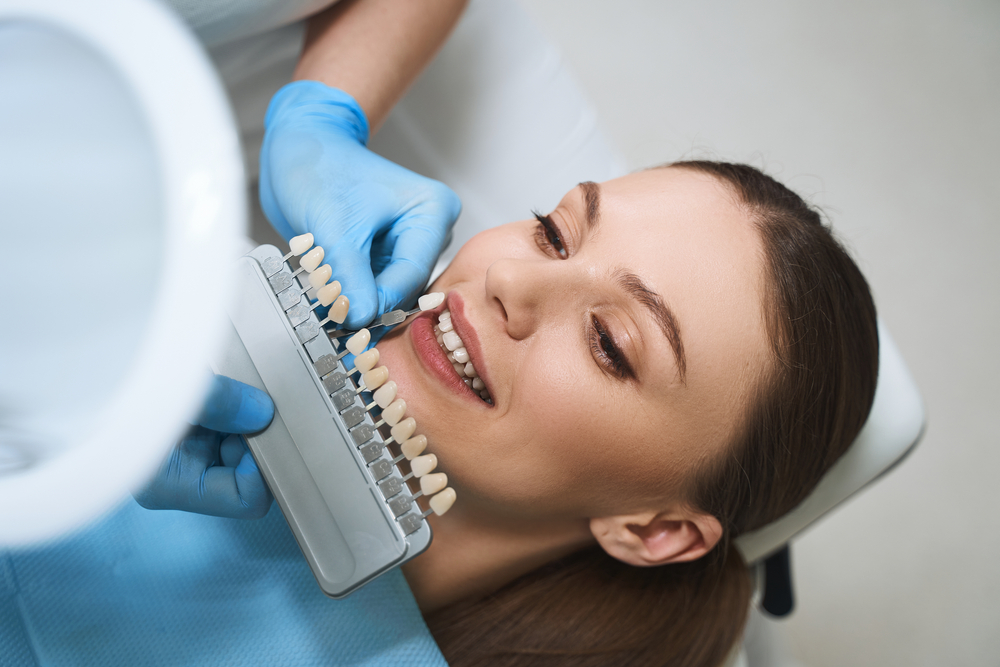 Cheerful young lady is lying in dental chair and choosing veeners colour with doctor help