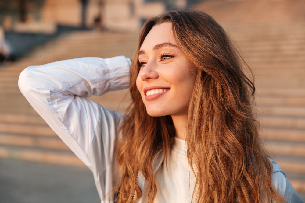 Woman with wavy hair smiling outdoors near a stone stairway 