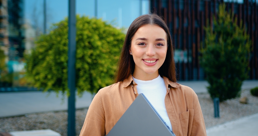 Close- up of beautiful brunette girl with long hair holding laptop in hands, looking at the camera with smiling against the background of modern building outdoors