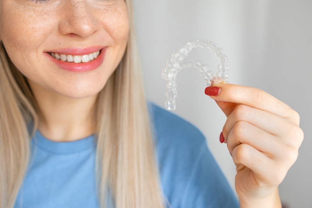 Close up young woman smiling with hand holding dental aligner retainer (invisible) isolated on white background for beautiful teeth