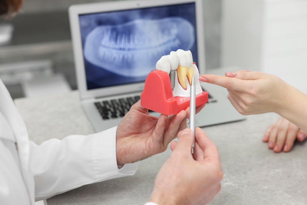 Understanding dental implants in Hampton Roads Dentist holding a dental implant model while a patient points to it, with a panoramic X-ray displayed on a laptop screen