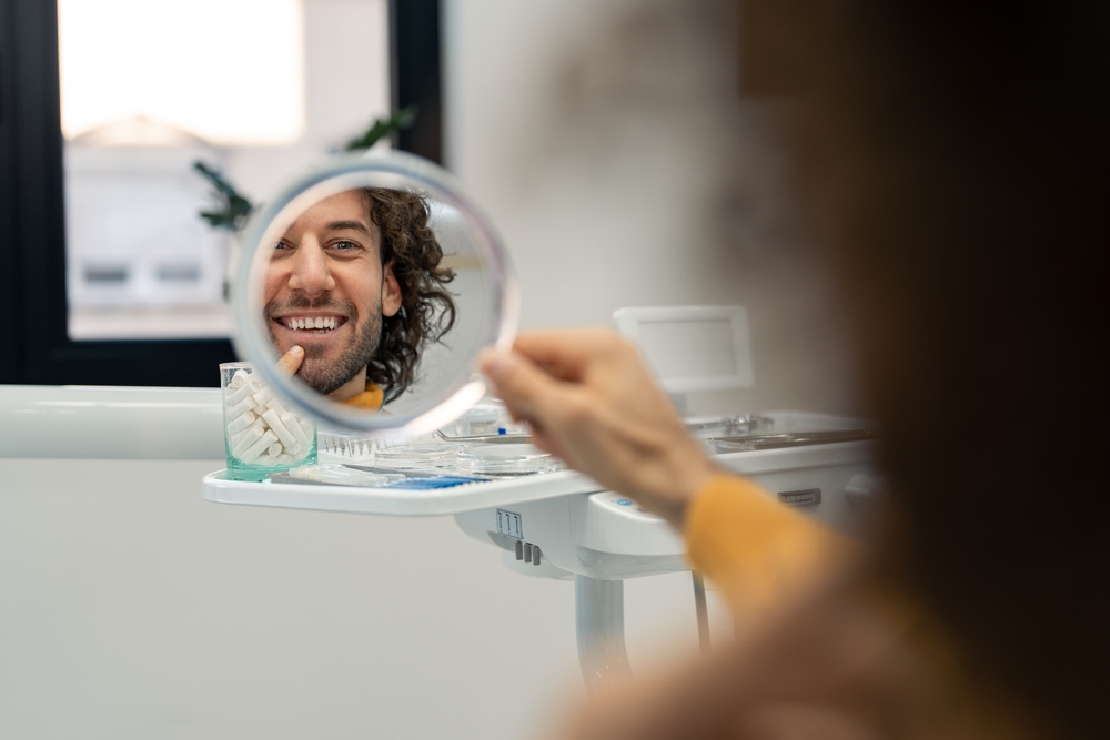 Handsome young man looking at mirror at dentist checking results after dental treatment teeth whitening new veneers feeling happy and satisfied, confident with healthy teeth smile.