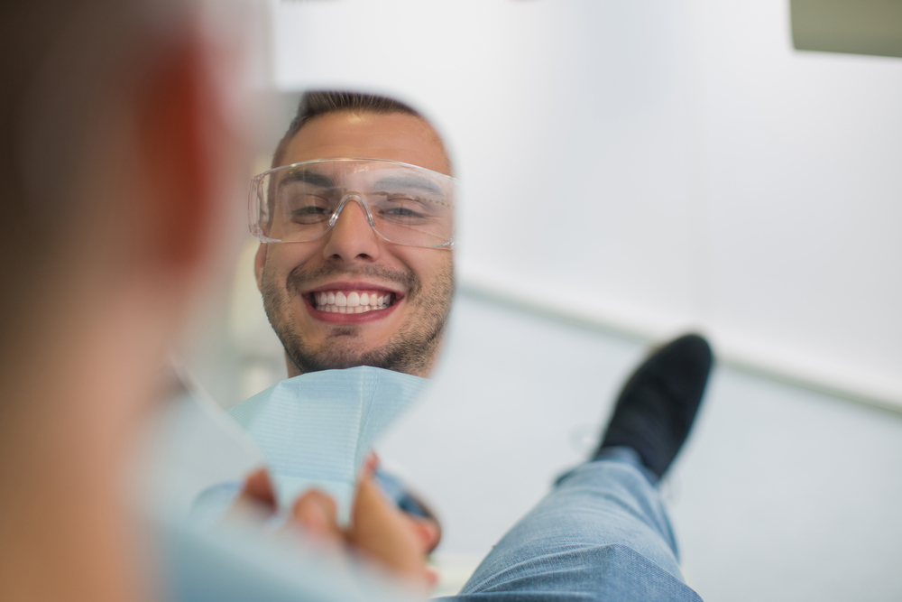 Happy patient looking in the mirror and smiling while sitting in dentist chair. Perfect smile.