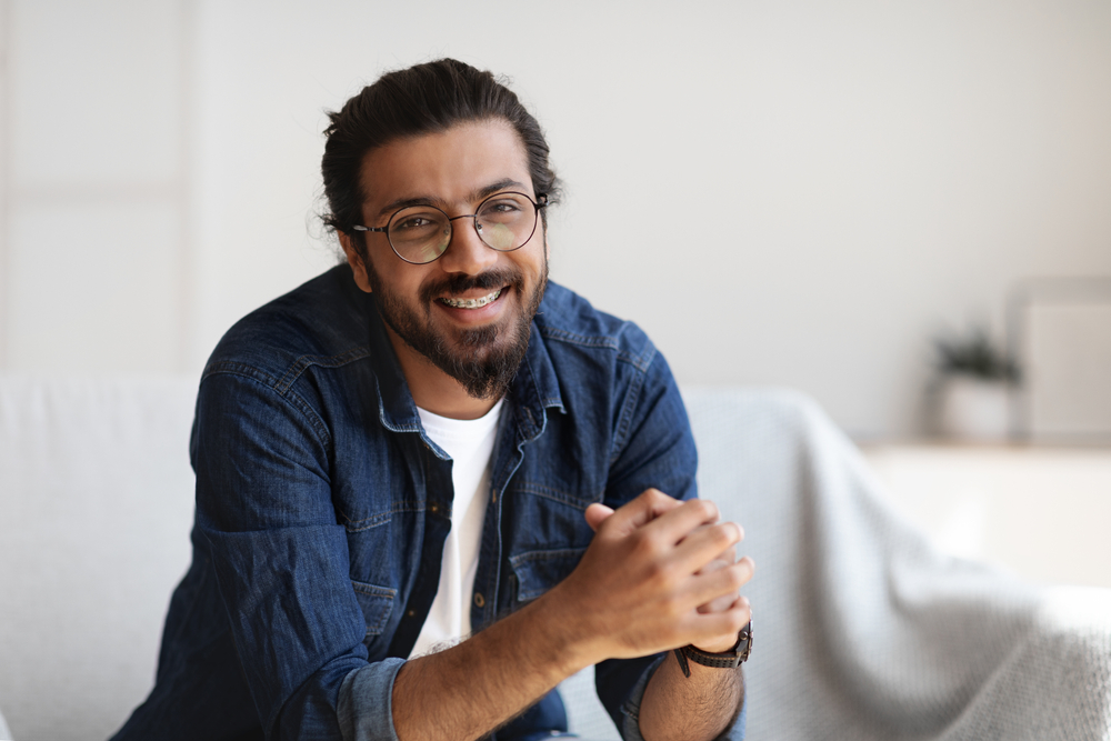 Man in a denim shirt smiling indoors with hands clasped 