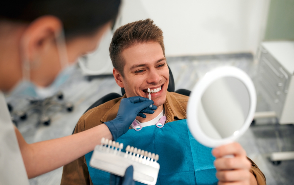Medicine, dentistry and healthcare concept - closeup of a dentist with tooth color samples choosing a shade for a male patient's teeth in a dental clinic looking at a mirror.
