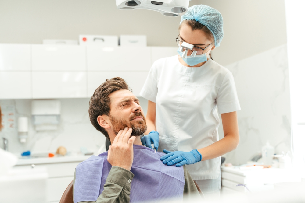 Man wincing in pain while a dentist in blue gloves examines his mouth