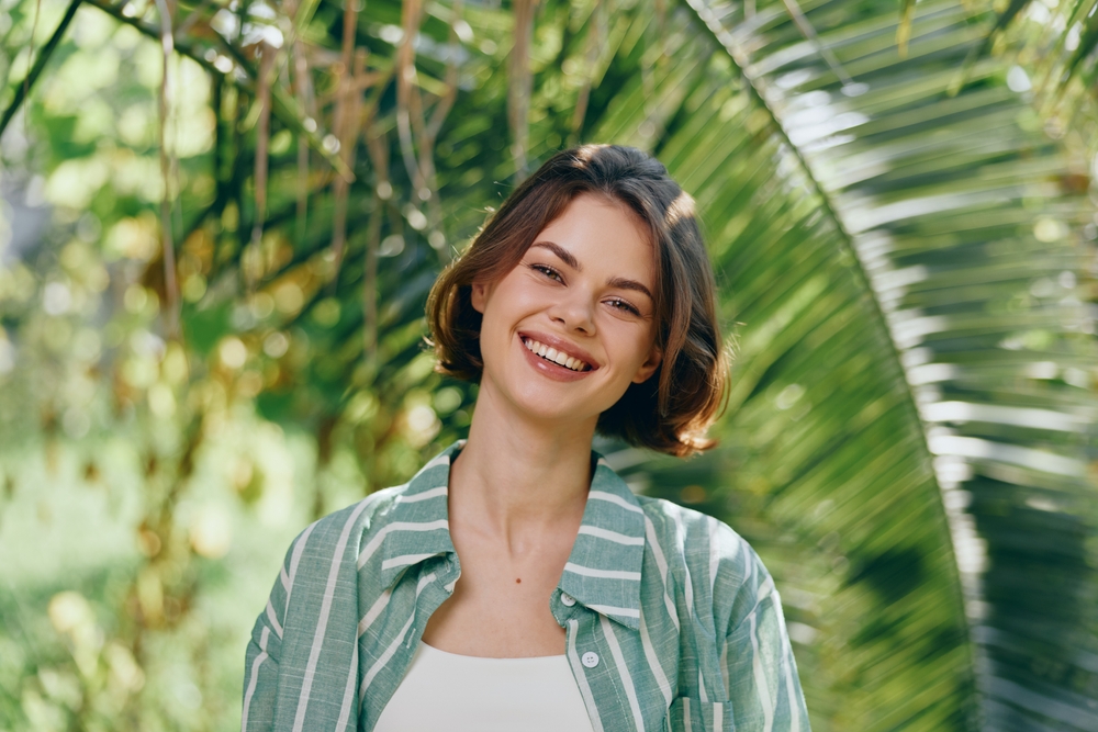 Smiling woman outdoors in natural light, wearing a striped green jacket over a white top, framed by lush tropical greenery and sunlit leaves.