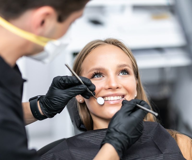 Dentist in black gloves examining a smiling patient's teeth with a mirror and explorer tool