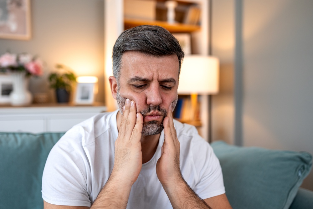 Toothache and dental pain. Adult man sitting on sofa holding painful jaw with hands. Healthcare and medicine concept. Handsome male suffering from sensitive teeth, gum inflammation or tooth decay.