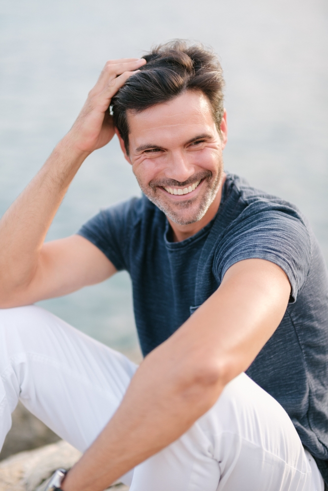 Man smiling outdoors near the water while running his hand through his hair