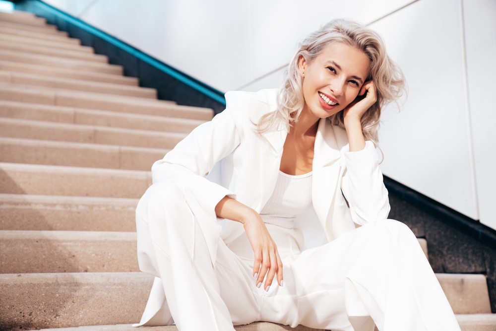 Young beautiful blond woman wearing nice trendy white suit jacket. Smiling model posing in the street at sunny day. Fashionable female outdoors. Cheerful and happy. Sits at the stairs 