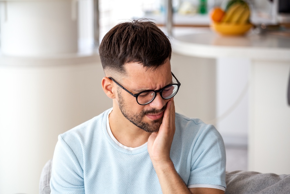 Young man in glasses suffering from severe toothache and jaw pain at home; concept for dental emergency, sensitive teeth, cavity, oral health, and need for professional dental care.