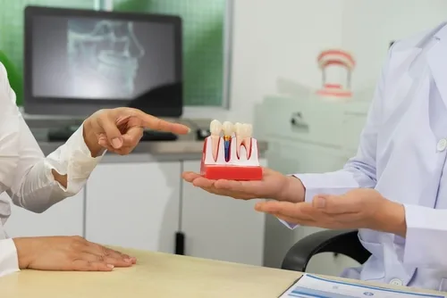 Discussing dental implant options in Hampton Roads Patient pointing to a dental implant model held by a dentist across a desk, with a panoramic X-ray on a monitor in the background