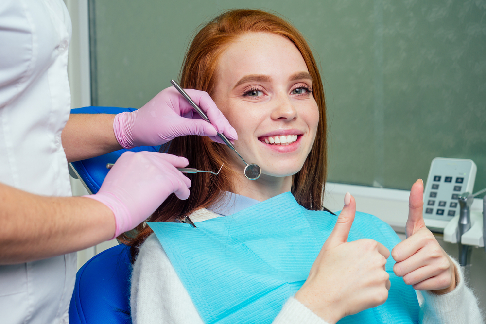Woman with red hair giving a thumbs up while a dentist examines her teeth with a mirror and explorer tool