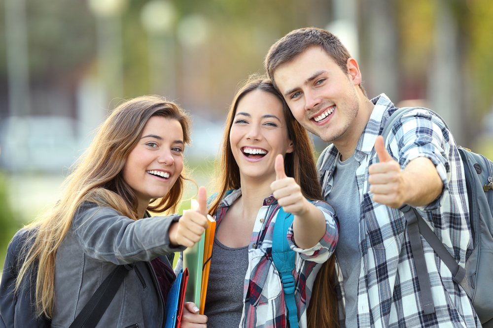 Three smiling students giving a thumbs up while holding backpacks and folders outdoors