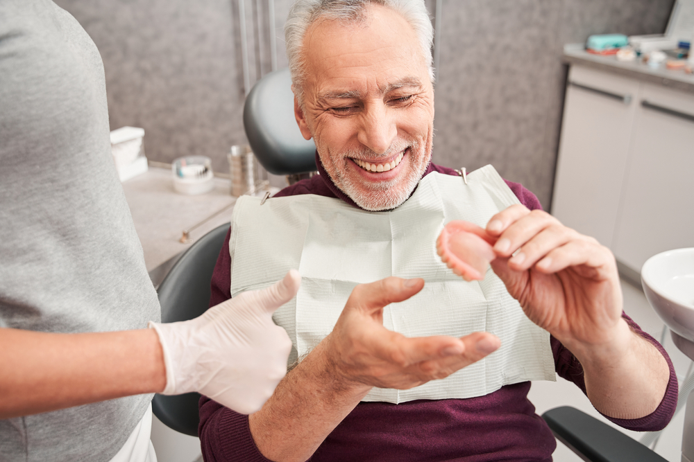 Custom dentures and partials in Norfolk Older man smiling while holding a denture in a dental chair