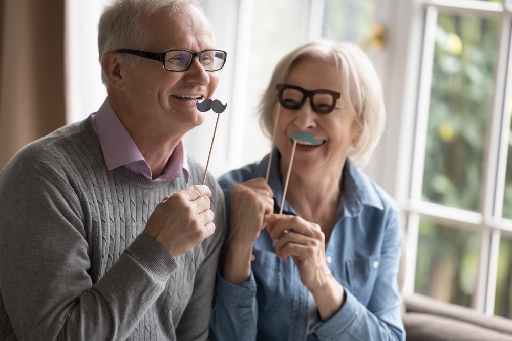 Enjoying life with confident dentures in Norfolk Elderly couple smiling while playfully holding mustache and glasses photo props