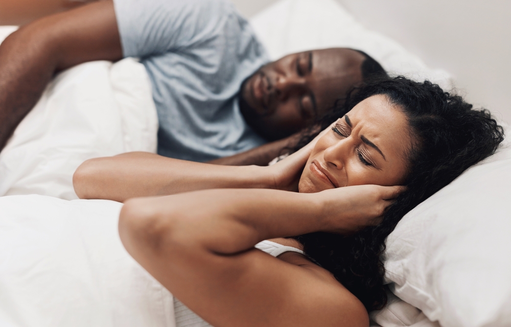 Woman covering her ears while lying next to a snoring partner, illustrating a common sleep apnea symptom in Hampton 