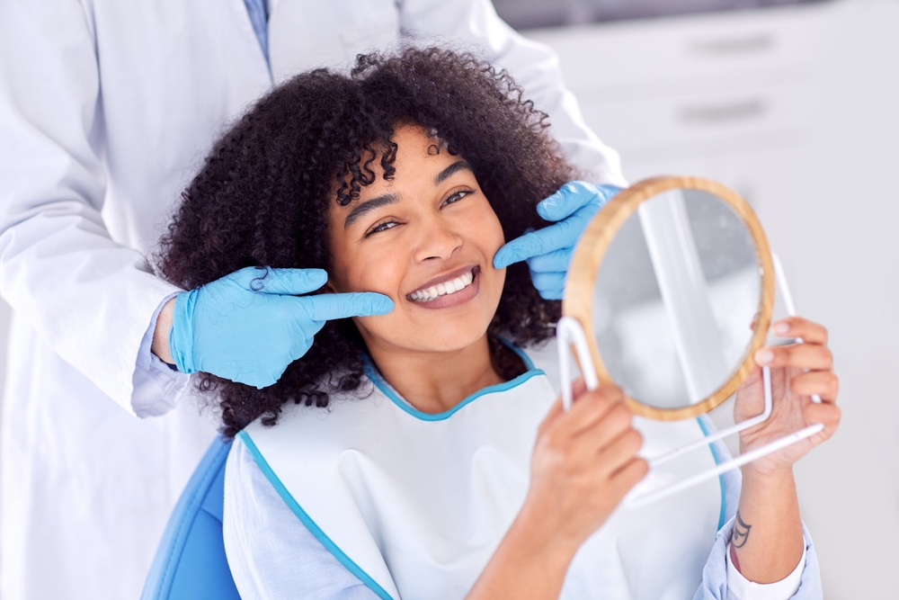 In-house dental lab smile results in Virginia Beach, VA, The Foleck Center Woman admiring her new smile in a hand mirror while a dentist highlights her custom restoration from The Foleck Center in-house dental lab, Virginia Beach, VA